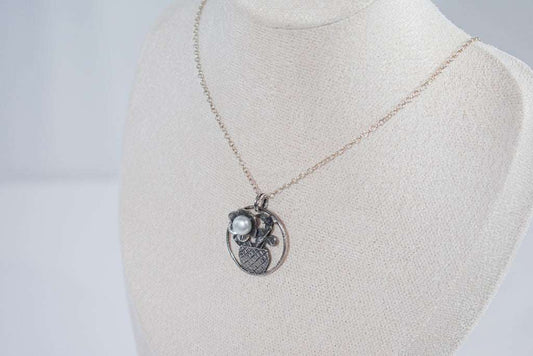 The Flower in the Basket pendant is a meticulously handcrafted antique silver masterpiece that exudes charm and elegance. Its intricate crisscross basket design cradles a delicate floral bloom, accented by a luminous pearl that adds a touch of sophistication.