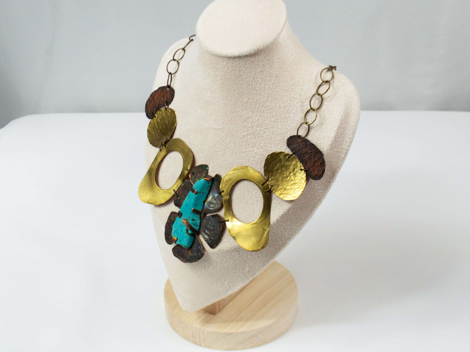 The necklace features a mix of brass and copper elements with hammered textures, creating an organic, artistic design. At the center is a striking 2-inch turquoise-dyed magnesite stone, irregularly shaped and set among darkened copper pieces. The necklace is finished with a brass chain made of large circular links, adding to its bold and rustic aesthetic. Perfect for witchy, bohemian, or office-chic styling.