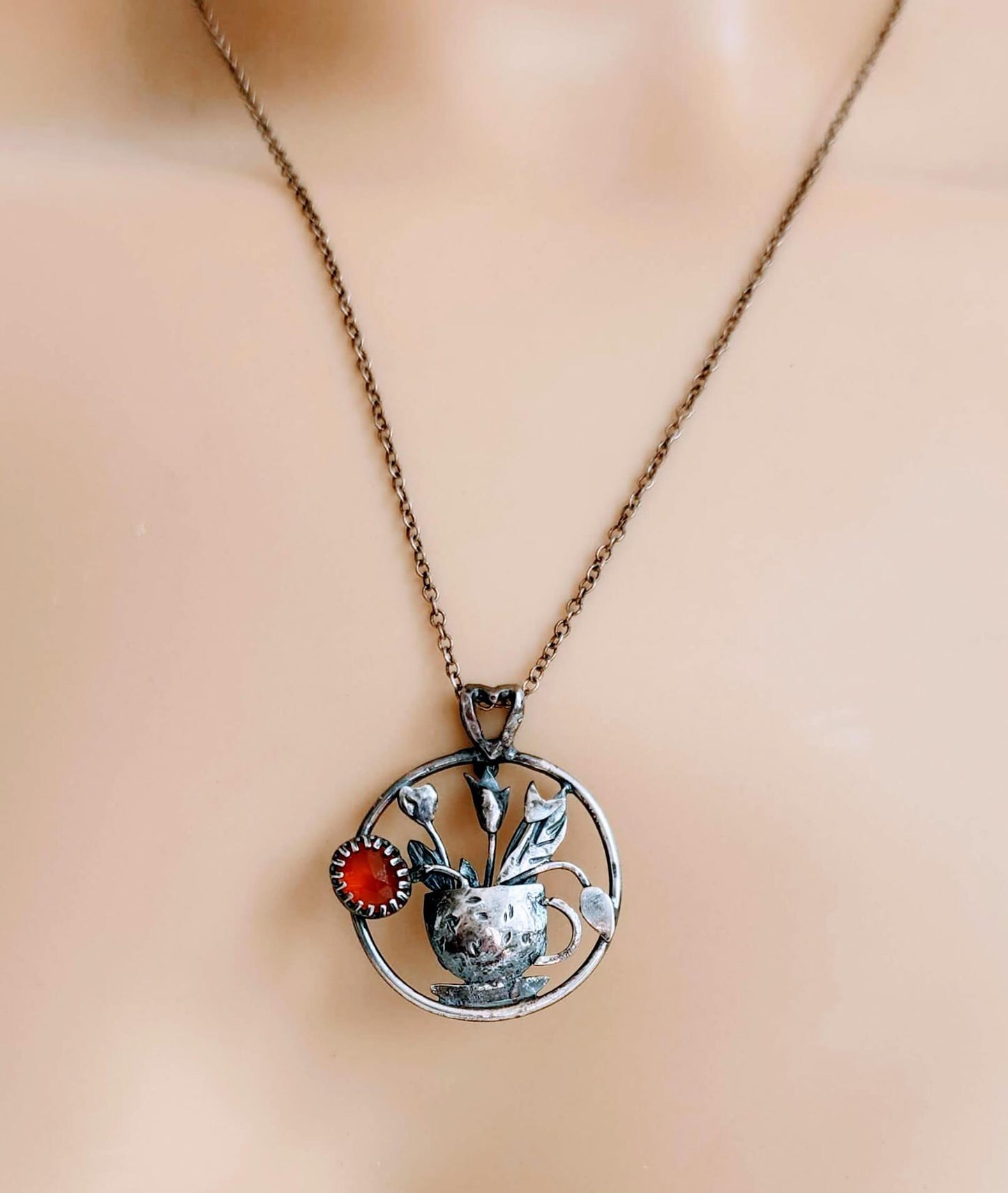Handcrafted sterling silver pendant necklace featuring a textured teacup with tulips and a glowing Carnelian gemstone, inspired by spring and tea rituals.