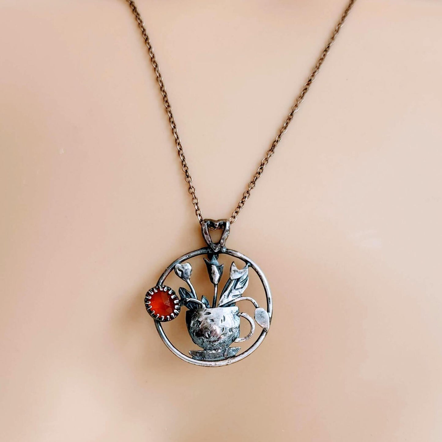 Handcrafted sterling silver pendant necklace featuring a textured teacup with tulips and a glowing Carnelian gemstone, inspired by spring and tea rituals.