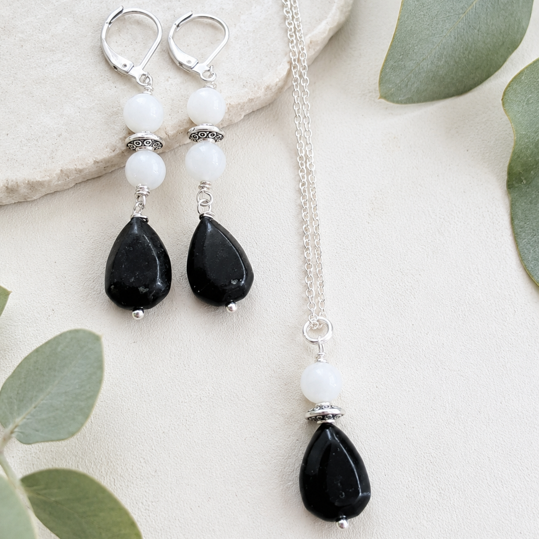 Black teardrop earrings and necklace with white beads on a light background with green leaves.