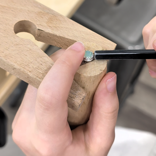 A close-up of hands holding a small ring in a wooden ring press while using a black tool to adjust or set the ring. The ring features a round light blue stone surrounded by a silver-toned halo.