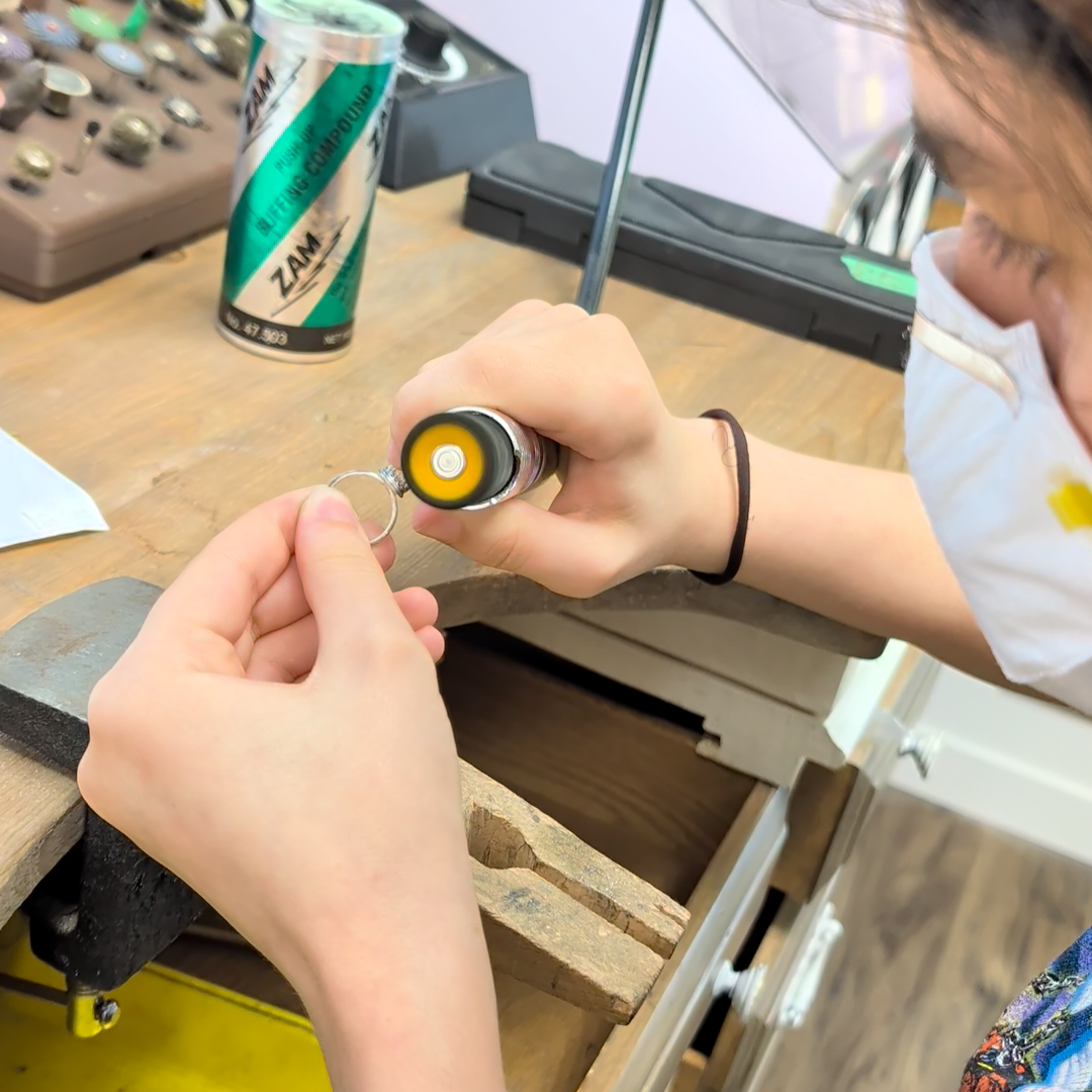 A close-up lifestyle shot of a person using a rotary tool with a small metal ring on a wooden workbench, surrounded by jewelry-making supplies.